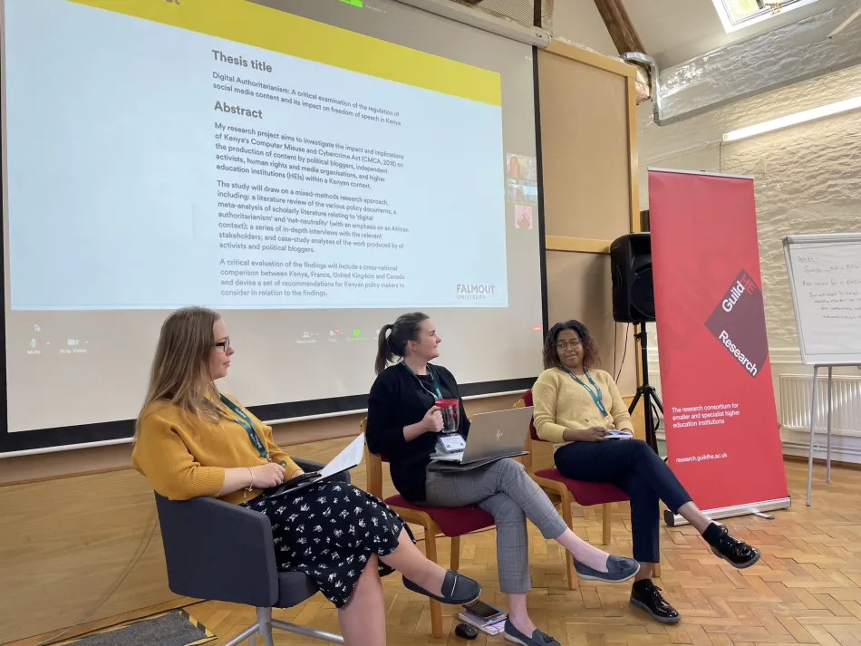  Three women seated as a panel talking about their experiences as PhD students, in front of a screen